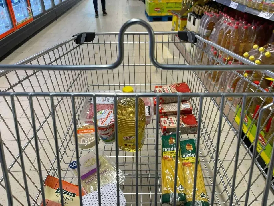 Un cărucior metalic conține sticle, cutii și alte produse ambalate, lângă rafturi cu băuturi și vitrine frigorifice.