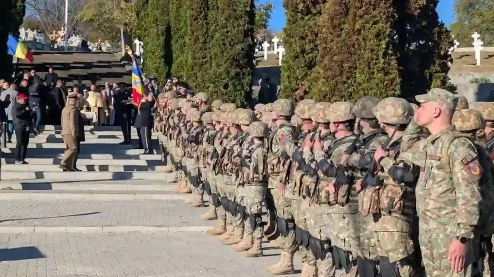 Militari în uniforme de camuflaj stau aliniați pe o alee; în fundal se văd alte persoane, un drapel și cruci albe.