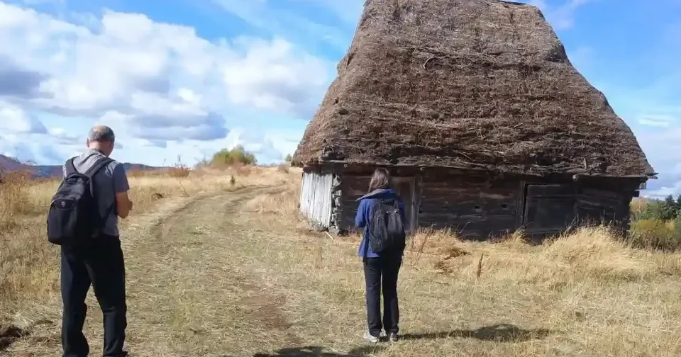 Două persoane cu rucsacuri stau pe o potecă lângă o construcție din lemn, cu acoperiș înalt din material vegetal.
