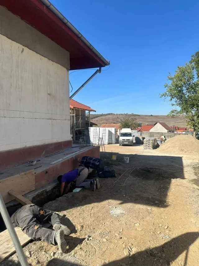 Două persoane se află lângă baza unei clădiri, una aplecată și cealaltă întinsă pe sol; în fundal se văd un vehicul și materiale de construcție.