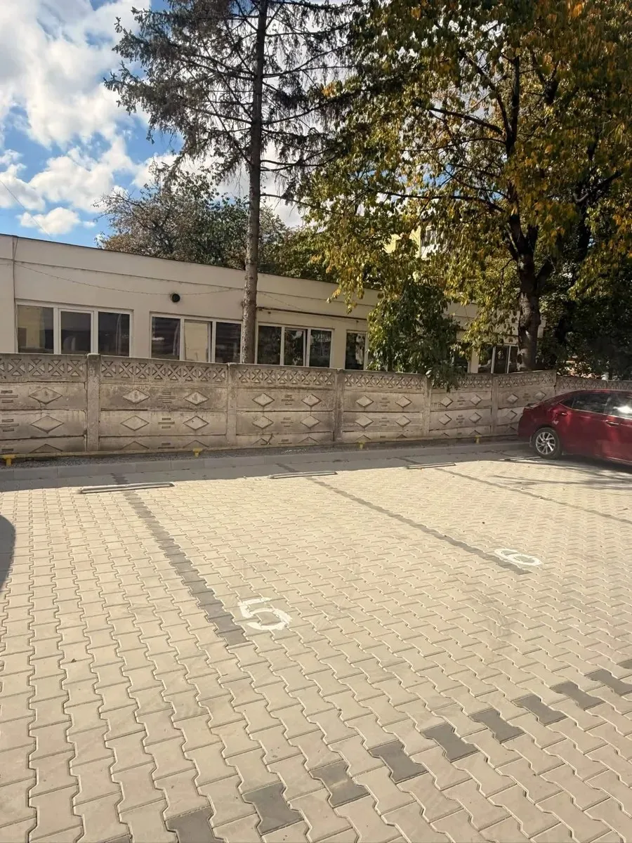 O parcare pavată cu dale deschise la culoare și marcaje albe se află în fața unei clădiri joase și a unui gard; în dreapta este o mașină roșie.