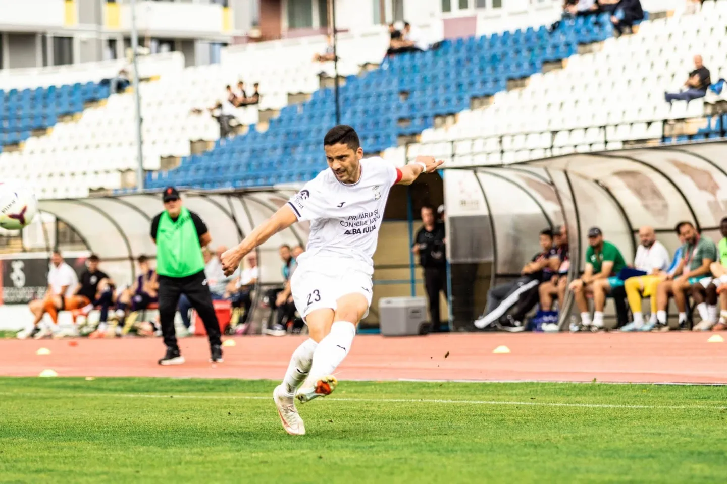 Un fotbalist în echipament alb lovește o minge pe gazon, iar în spate se văd persoane lângă bănci acoperite și tribune.