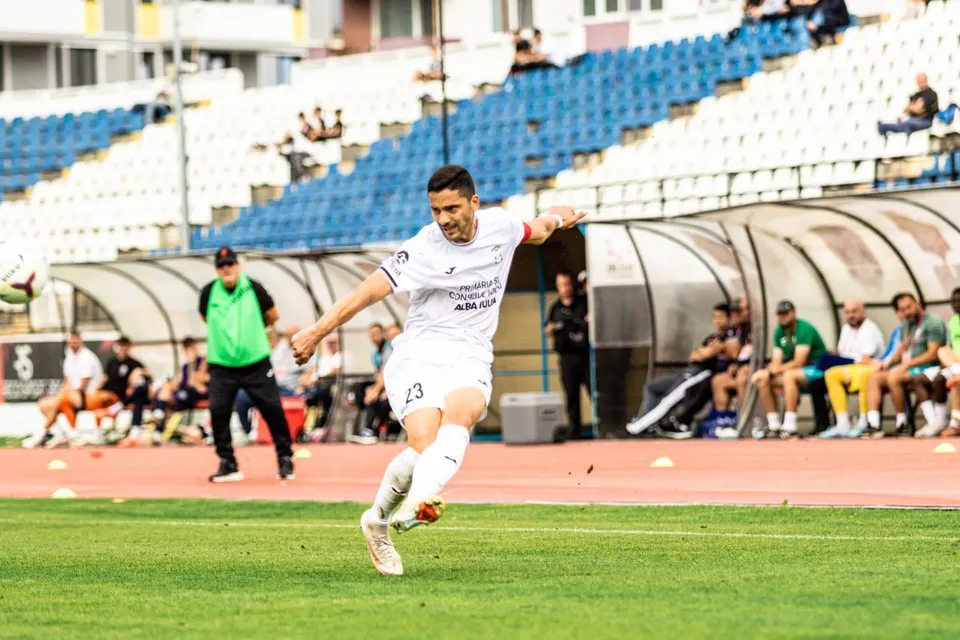 Un fotbalist în echipament alb lovește o minge pe gazon, iar în spate se văd persoane lângă bănci acoperite și tribune.