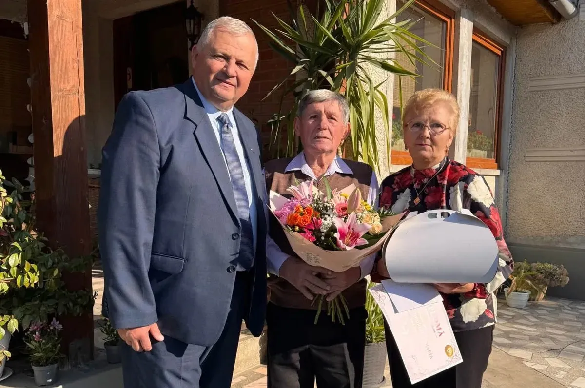 Un bărbat în costum stă lângă un bărbat și o femeie în vârstă; femeia ține un tort și o diplomă, iar bărbatul ține flori.