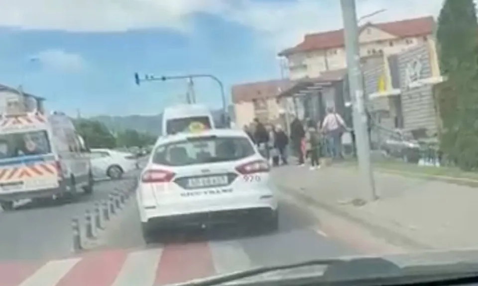 O ambulanță și mai multe autoturisme se află pe carosabil, iar un grup de persoane stă pe trotuar lângă o clădire.