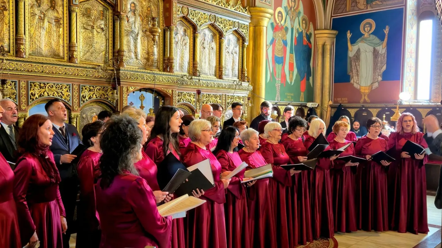 Un grup de persoane în robe vișinii ține mape negre și foi albe într-o sală cu pereți decorați și ferestre înalte.