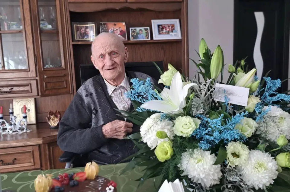 Un bărbat în vârstă, cu păr alb și sacou închis, stă într-o cameră, lângă un buchet mare de flori.