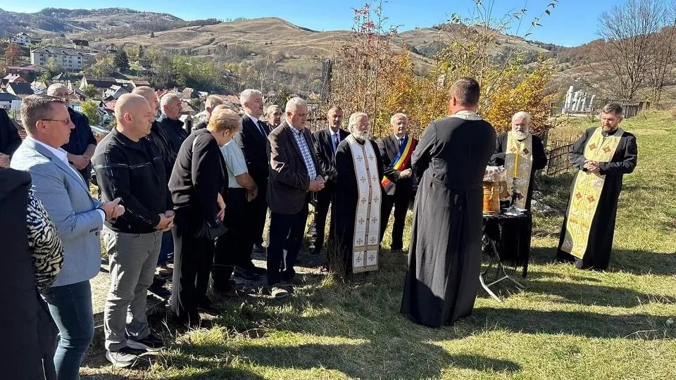 Mai multe persoane stau pe iarbă, în fața unor dealuri; trei poartă veșminte negre cu stole deschise la culoare, iar una ține o carte.