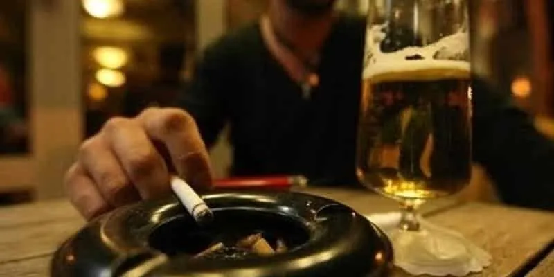În prim-plan se află o scrumieră cu un muc de țigară, lângă un pahar cu lichid auriu; în spate, o mână ține o țigară.