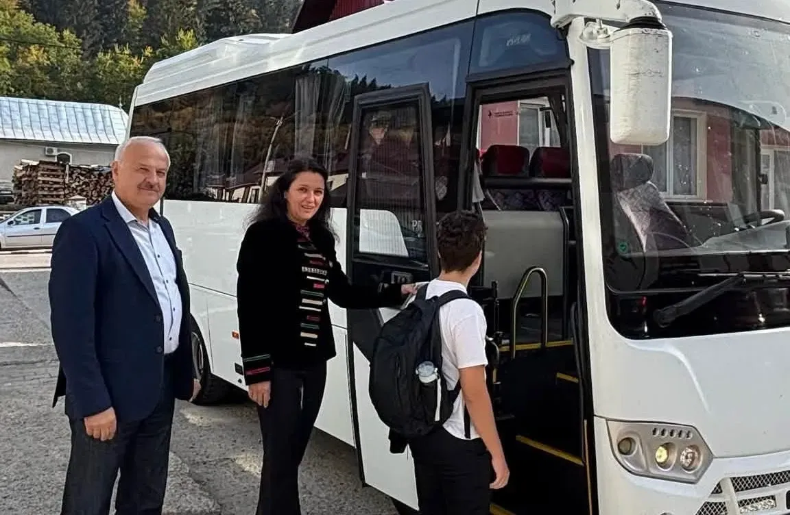 Un autobuz alb este oprit, cu ușa deschisă; un bărbat și o femeie stau lângă el, iar un elev cu ghiozdan se află în prag.