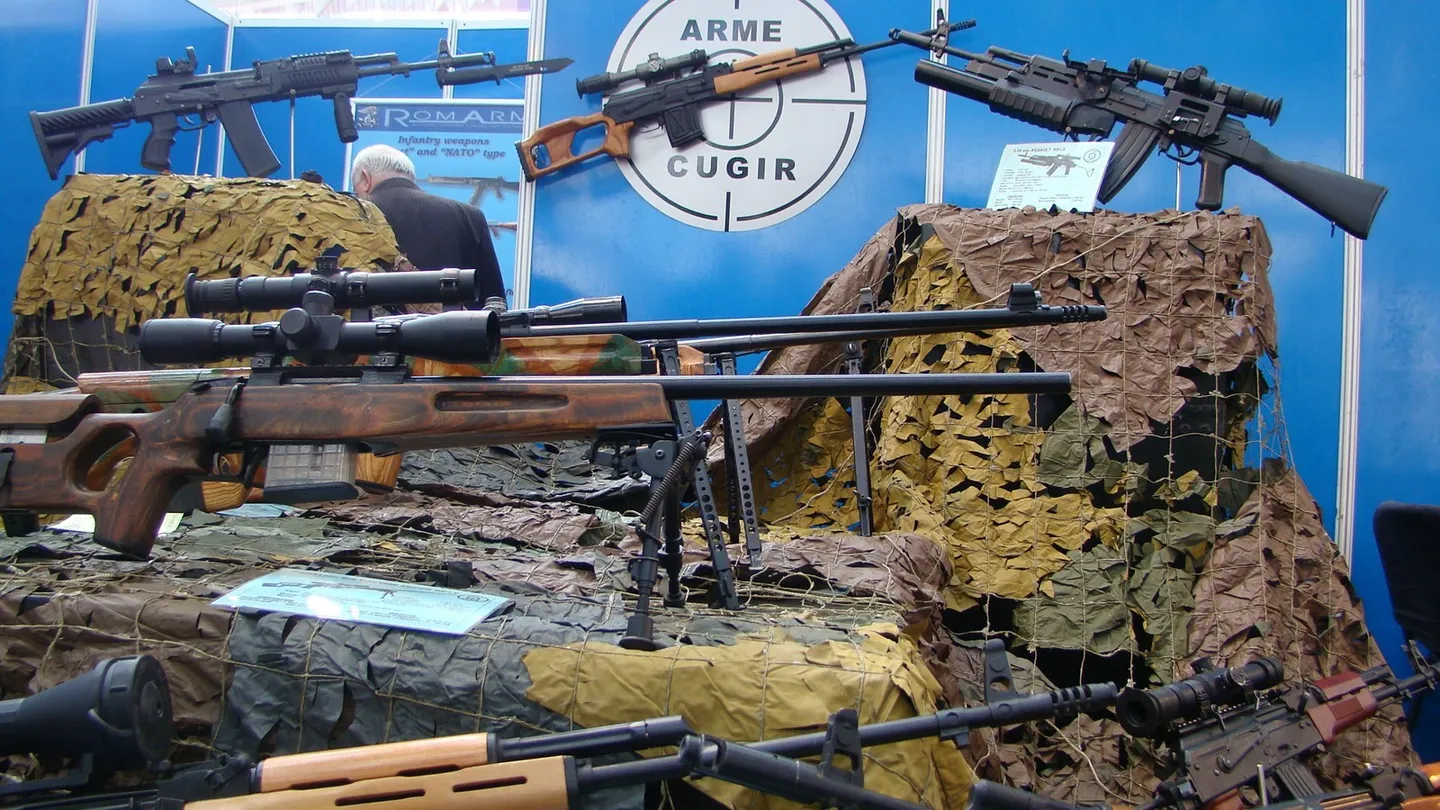 Mai multe arme sunt expuse pe suporturi, în fața unor panouri albastre și a unor materiale de camuflaj; în fundal apare o persoană.