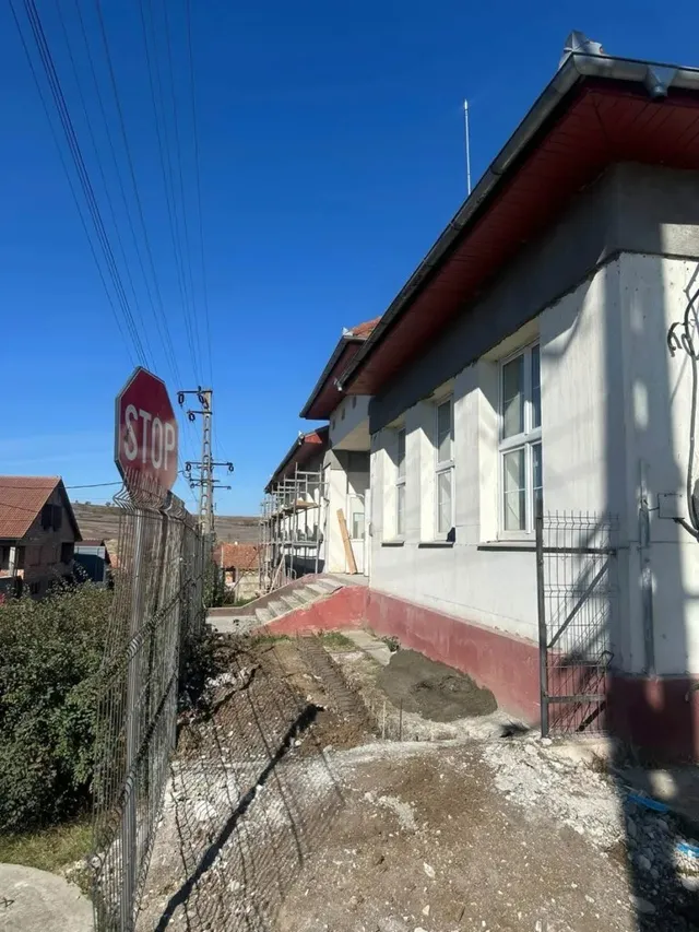 O clădire lungă, cu ferestre și acoperiș înclinat, se află lângă un gard metalic, iar schele și pământ răvășit sunt vizibile de-a lungul fațadei.