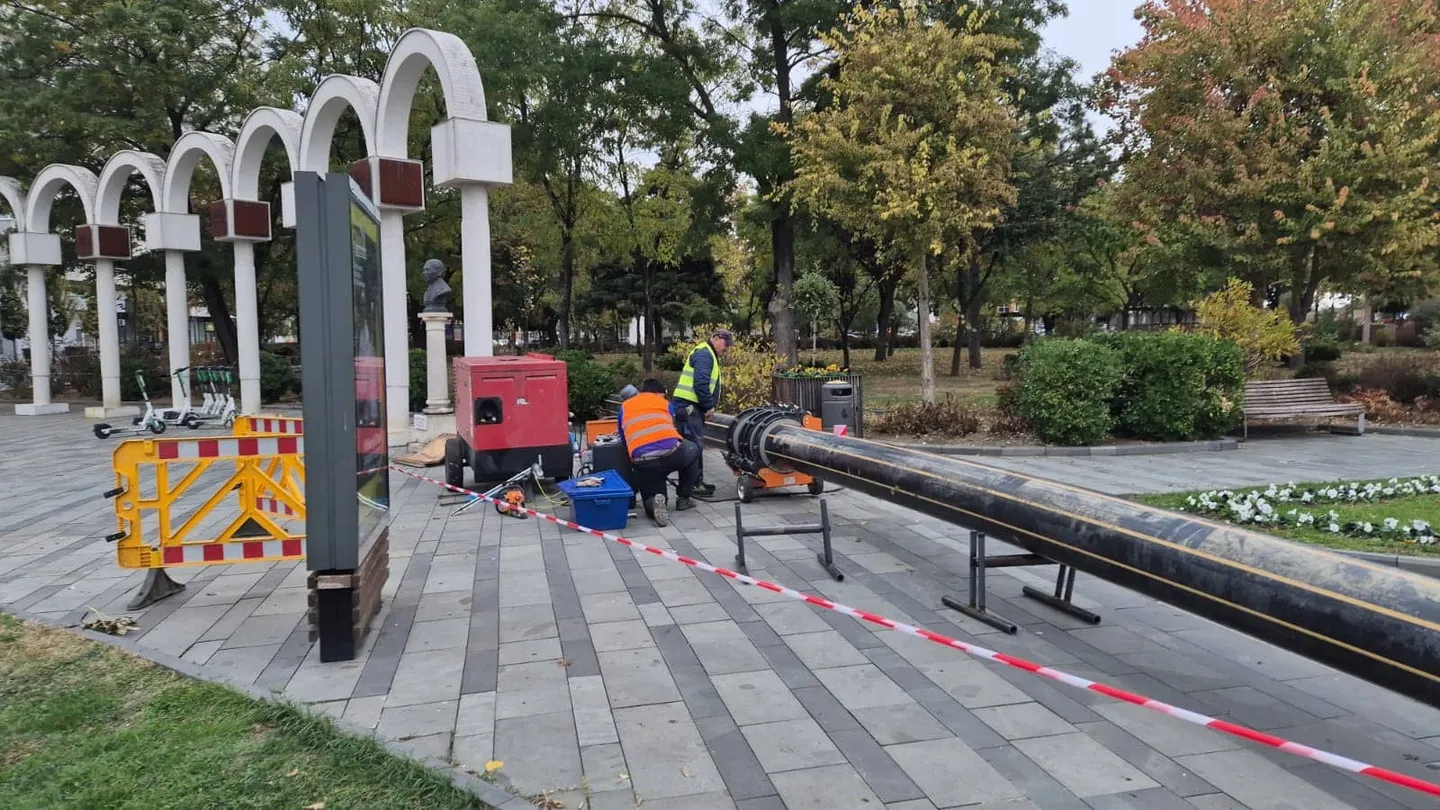 Doi muncitori cu veste reflectorizante se află lângă utilaje, conducte negre și bandă de delimitare, pe o alee cu dale.