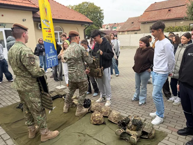 Doi bărbați în uniforme de camuflaj stau lângă mai mulți tineri, iar pe o prelată din prim-plan sunt așezate căști și echipamente militare.