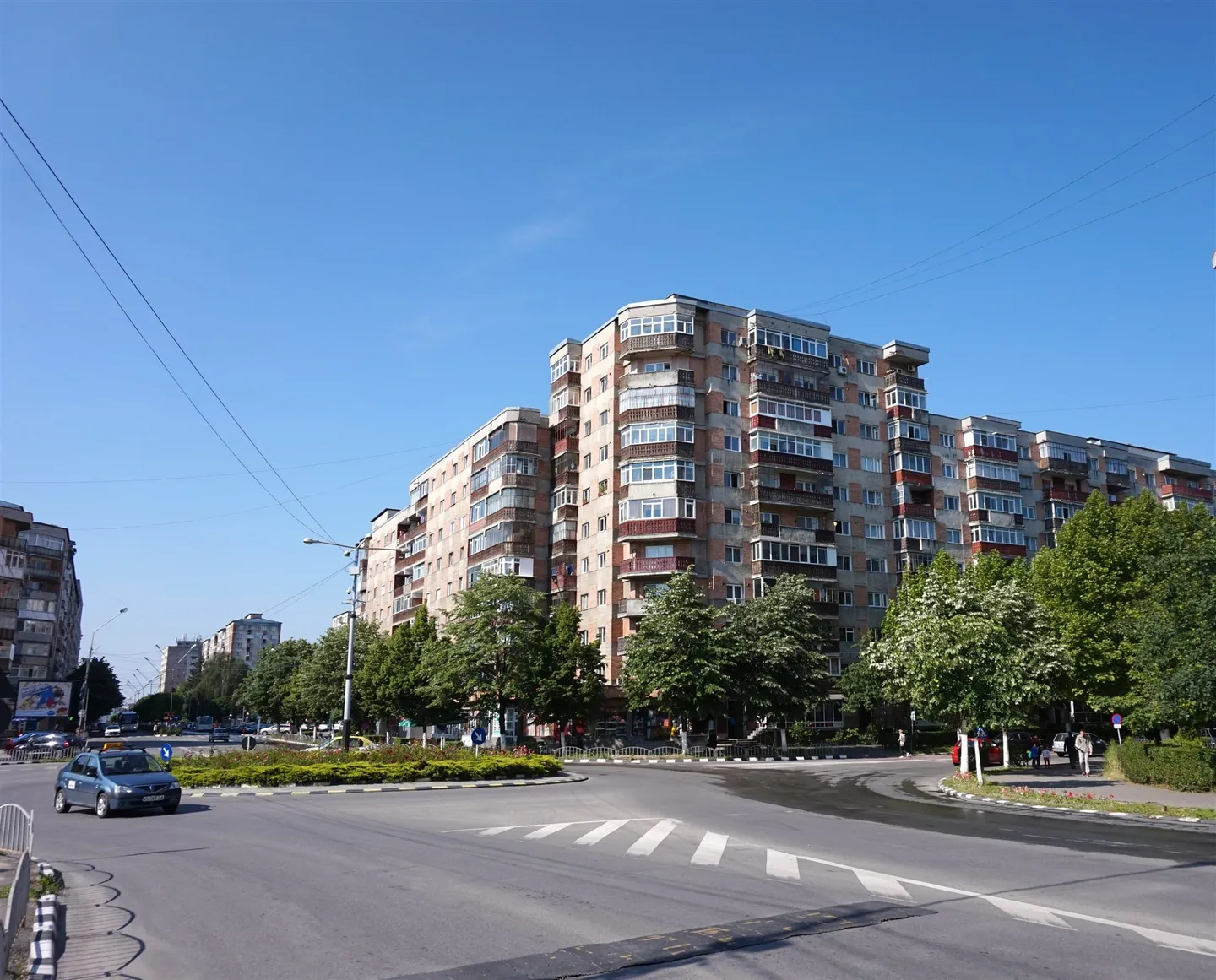 Blocuri înalte se află în spatele unor copaci, lângă o intersecție cu trecere de pietoni și un autoturism.