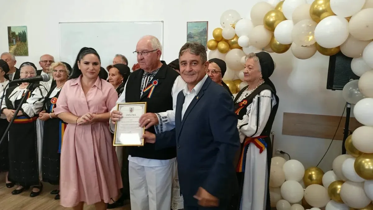 Mai multe persoane stau într-o încăpere decorată cu baloane albe și aurii, iar doi bărbați țin o diplomă înrămată.