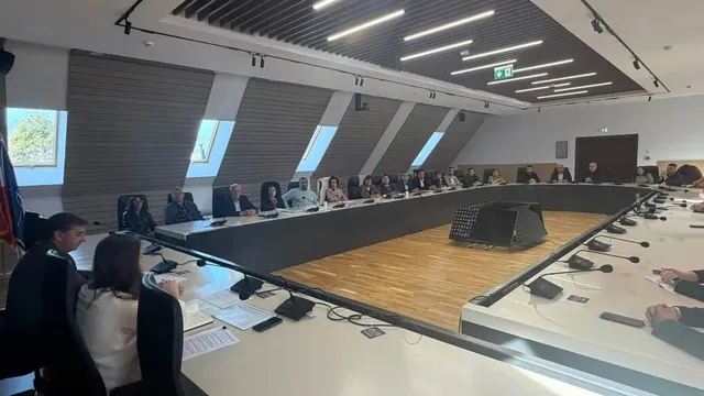 Mai multe persoane sunt așezate în jurul unei mese mari, cu documente, laptopuri și microfoane, într-o sală de conferințe.