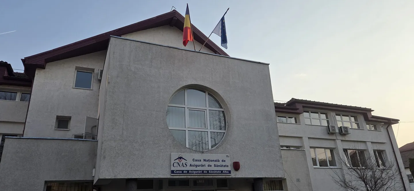 O clădire deschisă la culoare are o fereastră rotundă, două steaguri pe acoperiș și un panou cu text lângă intrare.