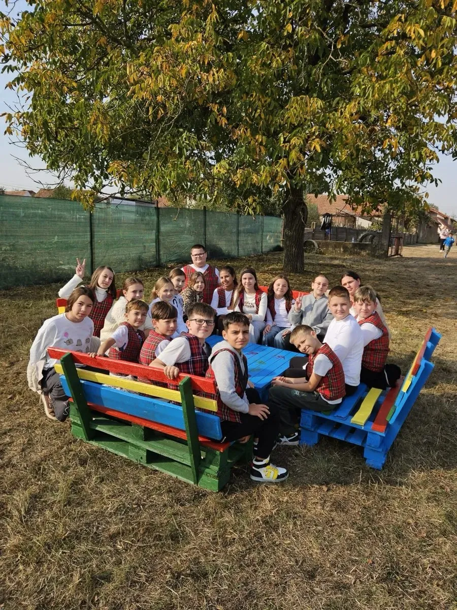 Un grup de copii stă în jurul unei mese albastre, pe bănci viu colorate, sub un copac cu frunze galbene.