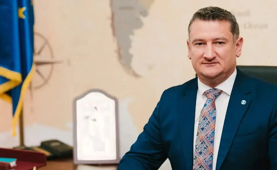 Un bărbat cu păr scurt poartă sacou bleumarin, cămașă albă și cravată cu model colorat; în spatele lui se văd un steag și o plachetă.