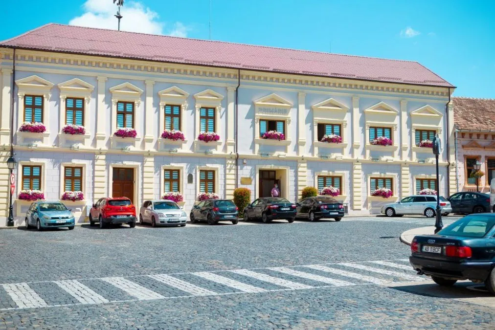 O clădire cu două niveluri, fațadă deschisă, acoperiș roșiatic și flori la ferestre se află lângă mașini parcate și o trecere de pietoni.