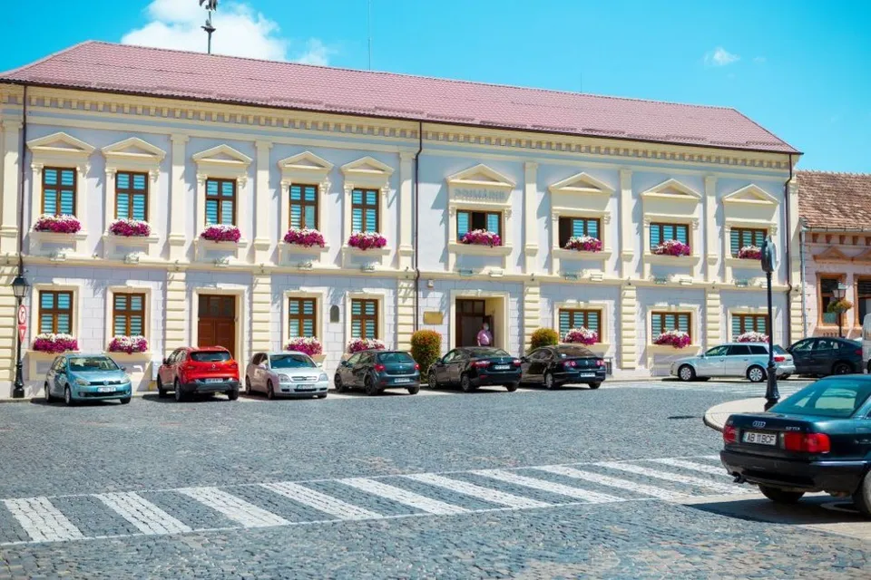 O clădire cu două niveluri, fațadă deschisă, acoperiș roșiatic și flori la ferestre se află lângă mașini parcate și o trecere de pietoni.