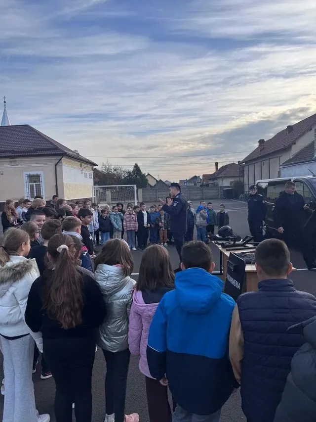 Un grup de copii stă în jurul unui bărbat în uniformă, lângă un vehicul închis la culoare; în fundal se văd clădiri.