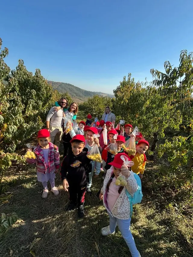 Un grup de copii, mulți purtând șepci roșii, stă între rânduri de pomi, cu câțiva adulți în spatele lor.