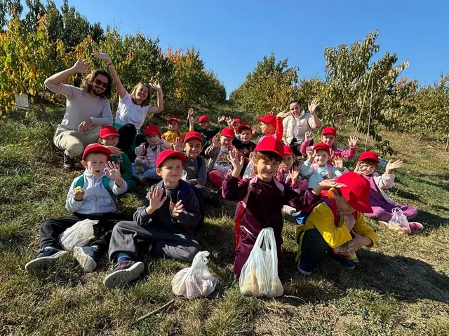 Un grup de copii, mulți purtând șepci roșii, stă pe iarbă; în spatele lor sunt doi adulți, iar în față se văd pungi și pomi.