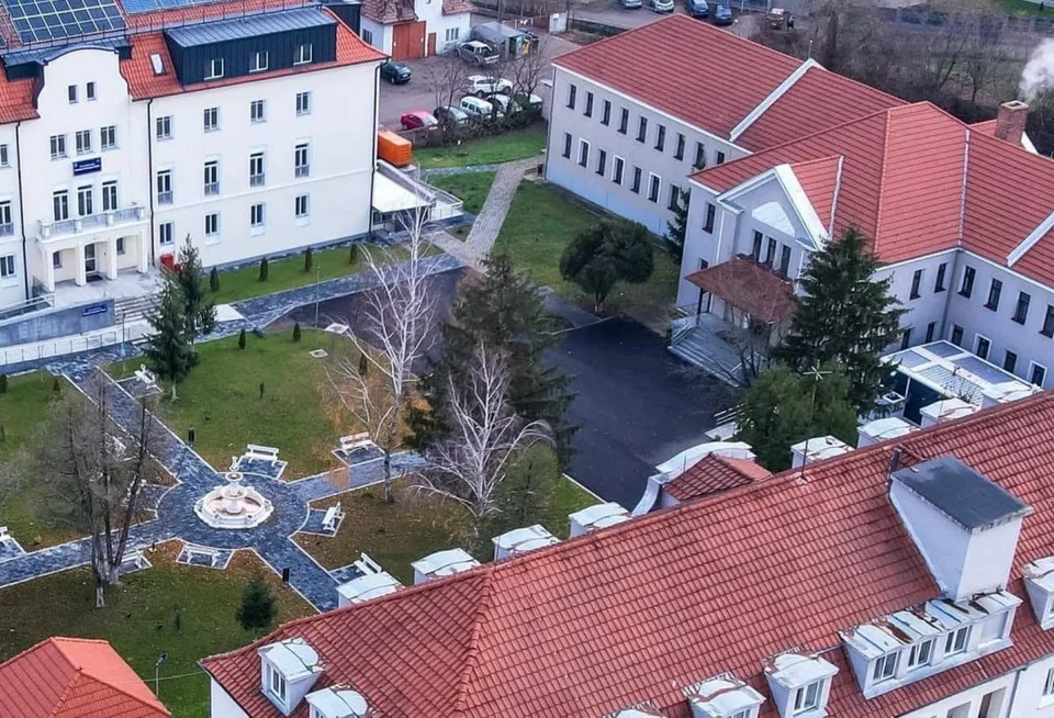 Mai multe clădiri albe cu acoperișuri roșii înconjoară o curte cu alei, copaci și câteva mașini parcate.