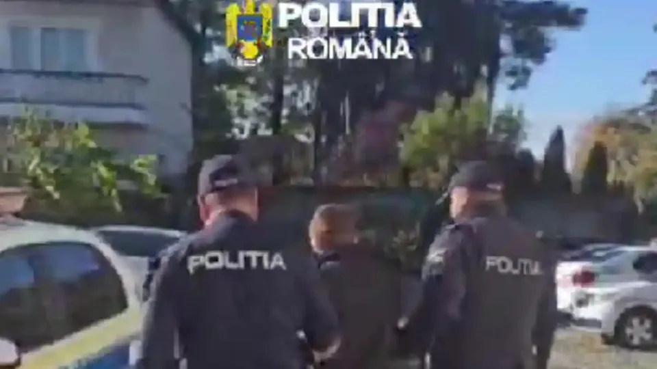 Trei persoane sunt văzute din spate lângă mașini parcate; două poartă jachete închise la culoare cu inscripția „POLIȚIA”.