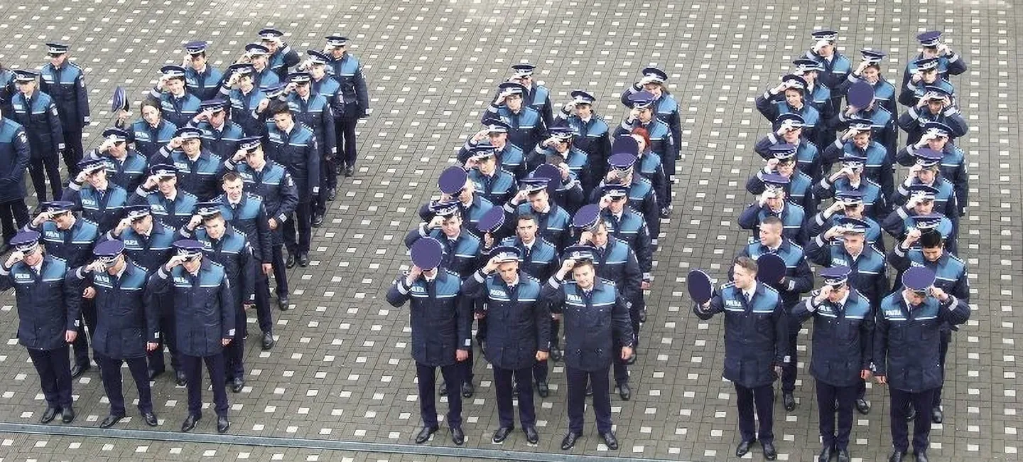 Numeroase persoane în uniforme bleumarin și șepci stau aliniate în grupuri pe o suprafață pavată, multe având mâna ridicată la tâmplă.