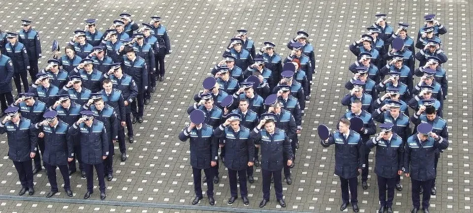 Numeroase persoane în uniforme bleumarin și șepci stau aliniate în grupuri pe o suprafață pavată, multe având mâna ridicată la tâmplă.