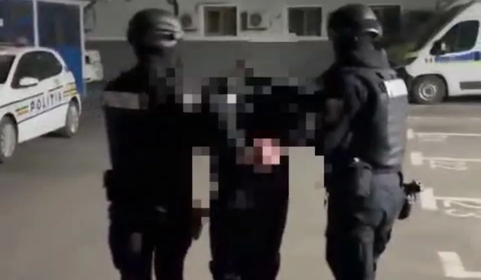 Două persoane îmbrăcate în uniforme închise la culoare stau de o parte și de alta a unei persoane cu fața pixelată; în spate sunt două autovehicule.