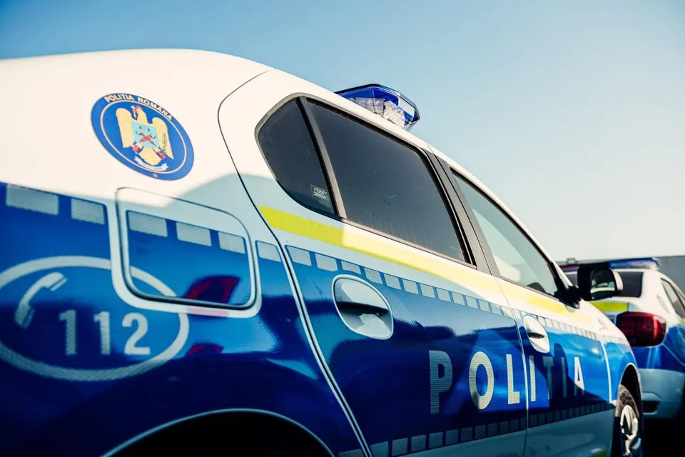 Un autoturism alb-albastru inscripționat „POLIȚIA”, cu numărul 112, emblemă și girofar, este văzut lateral, lângă un alt vehicul.