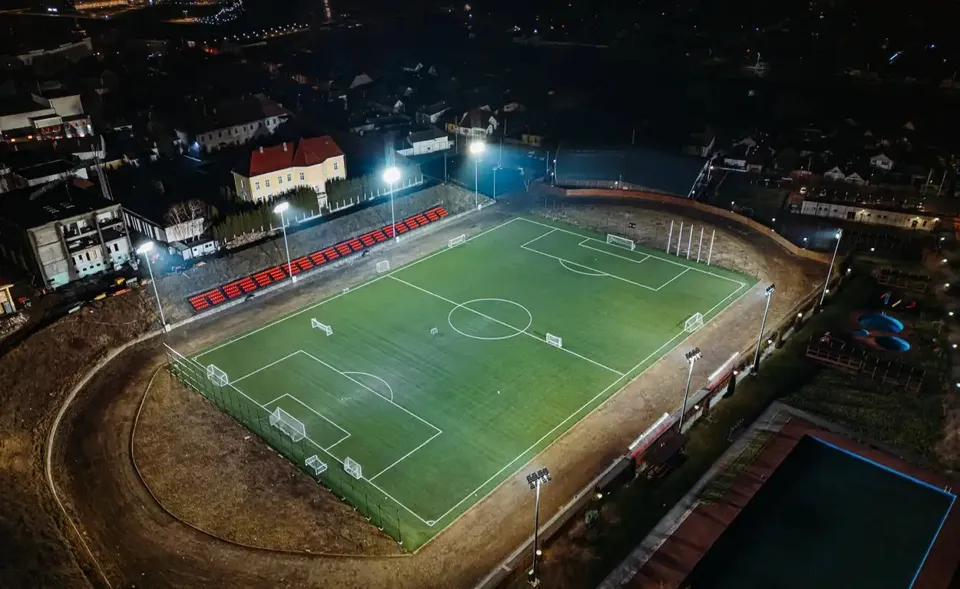 Un teren verde cu marcaje albe și porți este înconjurat de o pistă, tribune cu scaune roșii, stâlpi iluminați și clădiri.