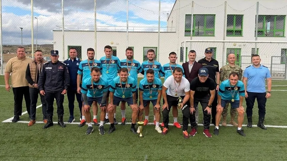 Un grup de bărbați pozează pe un teren cu gazon artificial; unii stau în picioare, iar alții se apleacă, purtând tricouri sportive.