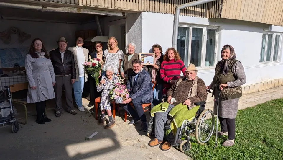 Mai multe persoane stau în fața unei clădiri; două persoane vârstnice sunt așezate, una într-un scaun cu rotile, iar în grup se văd flori și un obiect înrămat.
