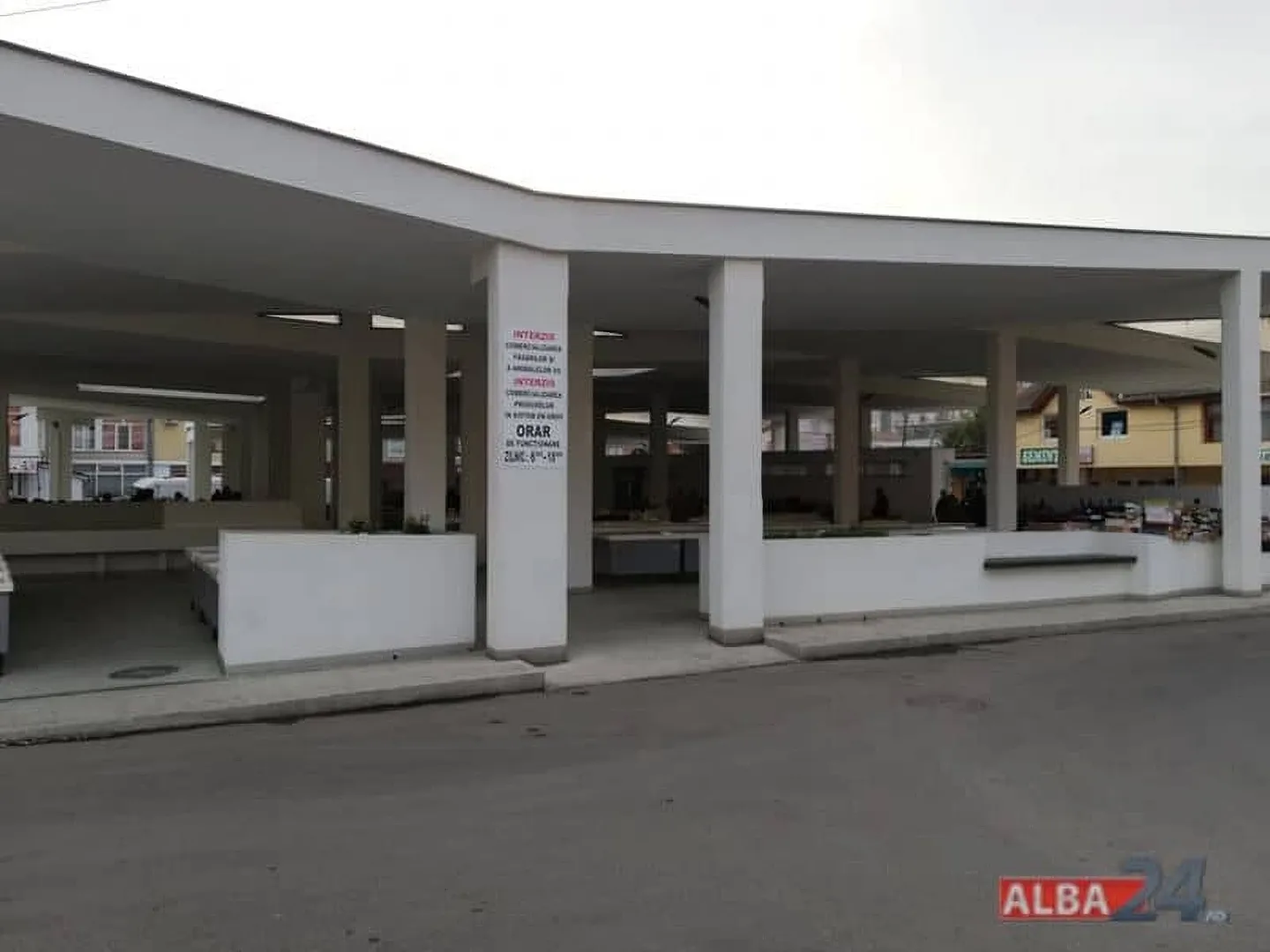 O construcție acoperită, cu stâlpi albi și spații deschise, se află dincolo de un carosabil; pe un stâlp este montat un panou.