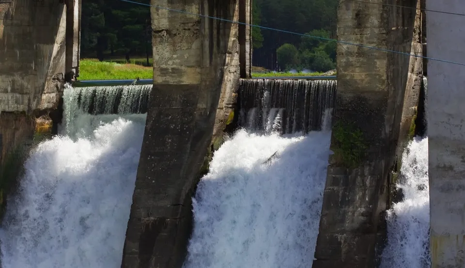 Apa curge în cascade între piloni masivi din beton, cu vegetație verde vizibilă în fundal.