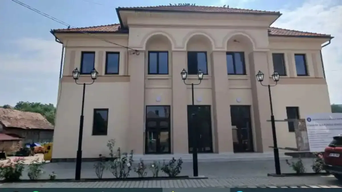 O clădire cu două niveluri, fațadă deschisă și ferestre întunecate se află în spatele unei alei pavate, cu felinare și o mașină roșie în dreapta.