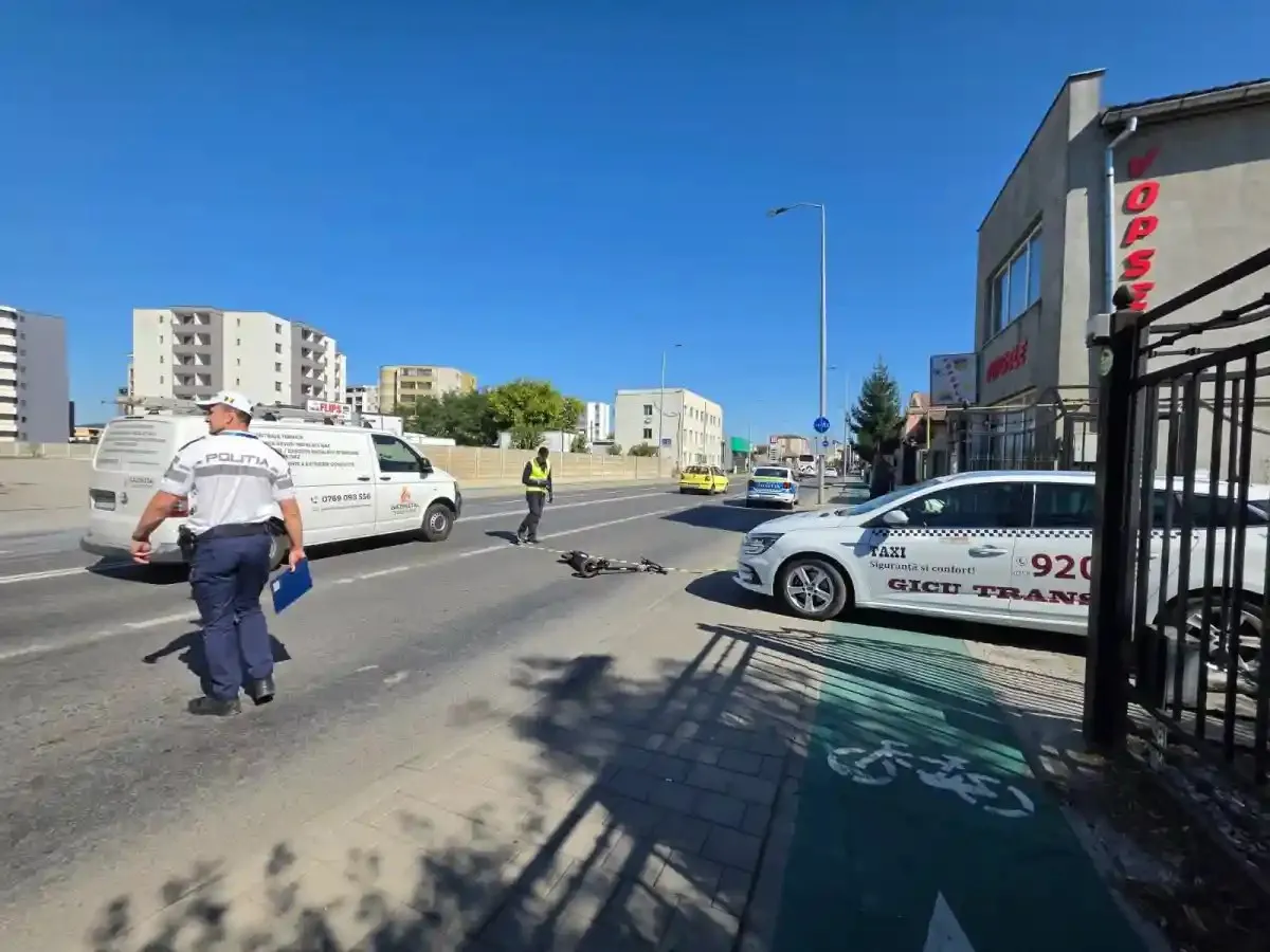 Pe carosabil se află o trotinetă culcată, două persoane, o autoutilitară albă și mai multe autoturisme, lângă clădiri și un gard metalic.