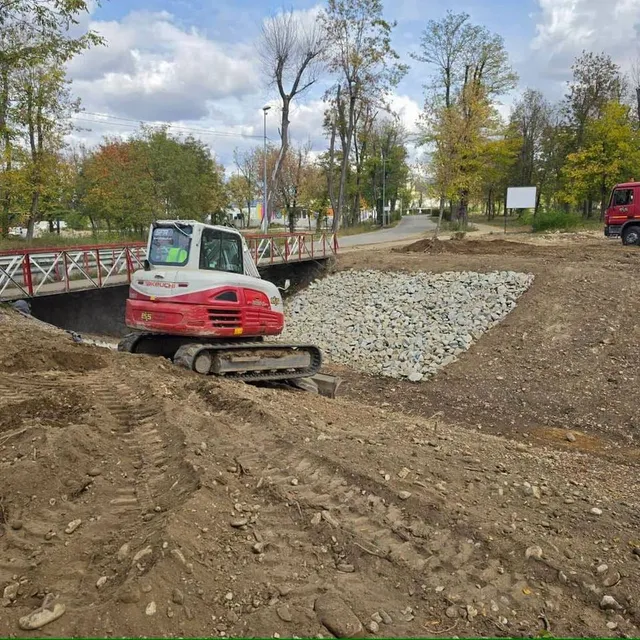 Un excavator roșu și alb se află pe un teren cu pământ răscolit, lângă pietriș, o pasarelă, copaci și un camion roșu.