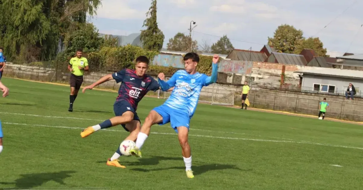 Doi jucători de fotbal, unul în echipament albastru și celălalt în echipament bleumarin, se dispută mingea pe un teren verde.