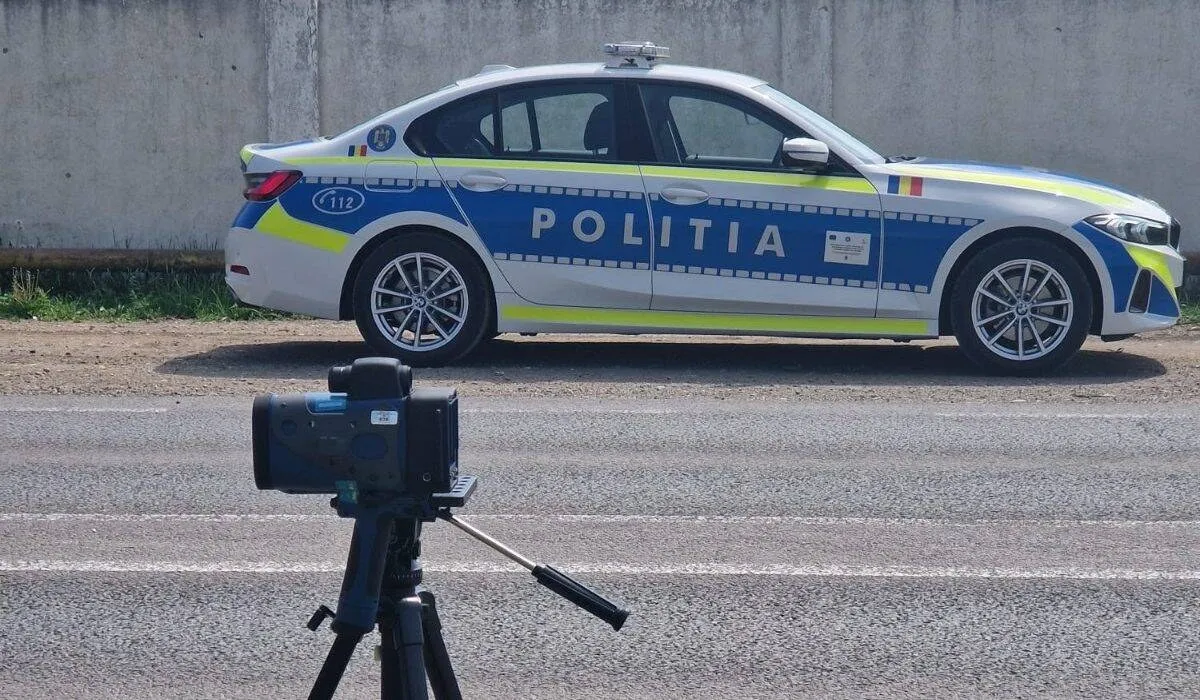 Un aparat negru montat pe trepied se află în prim-plan, iar în spate este parcată o mașină de poliție lângă un zid.