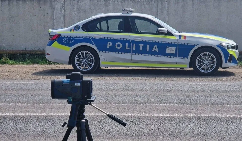 Un aparat negru montat pe trepied se află în prim-plan, iar în spate este parcată o mașină de poliție lângă un zid.