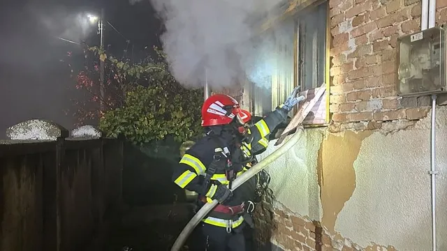 Un pompier cu cască roșie și uniformă neagră cu benzi reflectorizante ține un furtun lângă o fereastră, din care iese fum.
