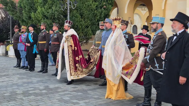 Două persoane cu coroane și mantii lungi stau în fața unei clădiri cu arcade, alături de persoane în uniforme și haine închise la culoare.