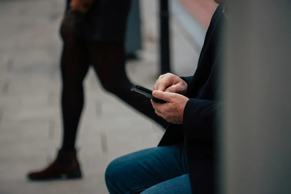 O persoană așezată, în pantaloni albaștri și jachetă închisă la culoare, ține un telefon; în fundal se vede o persoană în mers.