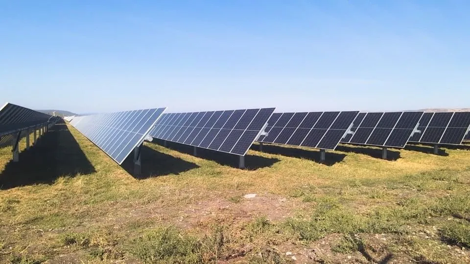 Rânduri de panouri solare înclinate se întind pe un teren acoperit cu iarbă rară, sub un cer senin.